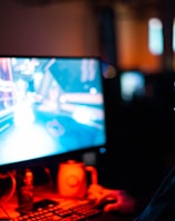 A person wearing headphones is sitting in front of a computer monitor. The screen displays a vibrant, illuminated video game scene, while the person appears focused on gaming. The ambient lighting is dark with red and blue tones, and a backlight from the monitor highlights the headphones.