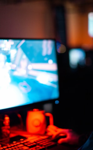 A person wearing headphones is sitting in front of a computer monitor. The screen displays a vibrant, illuminated video game scene, while the person appears focused on gaming. The ambient lighting is dark with red and blue tones, and a backlight from the monitor highlights the headphones.