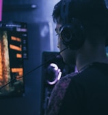 A person wearing headphones is engaged in gaming on a computer, with a large screen displaying a video game scene, showing a wooden building and a path.