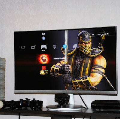 A flat-screen TV displays a video game interface featuring a character from Mortal Kombat in the center. Beside the TV is a stack of video game cases for the PlayStation 3, including titles like FIFA 14 and Grand Theft Auto. Below the TV, there is a gaming console, controllers, and other gaming equipment.