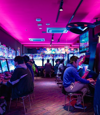 A vibrant arcade with rows of gaming machines adorned with colorful screens. Several people are immersed in playing games, seated on stools in a dimly lit room accented by an intense pink ceiling light. The atmosphere is lively and engaging.