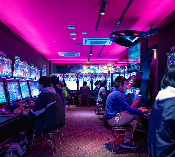 A vibrant arcade with rows of gaming machines adorned with colorful screens. Several people are immersed in playing games, seated on stools in a dimly lit room accented by an intense pink ceiling light. The atmosphere is lively and engaging.