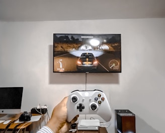 A person is holding a gaming controller in front of a flat-screen television displaying a racing video game. The room has a computer setup on a wooden desk with cables, a desktop computer monitor, and other accessories. The focus is on the interaction between the person and the gaming environment.
