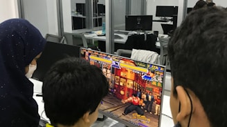 A group of children is gathered around a desktop computer, engaged in playing a colorful, action-packed arcade game displayed on the monitor. The setting appears to be a modern, well-lit office or gaming space with large glass windows, showing multiple computer workstations in the background.
