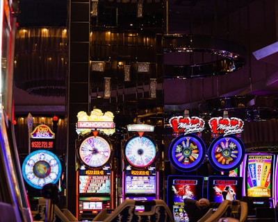 A lively casino scene featuring a row of bright, colorful slot machines with themes like Monopoly, Michael Jackson, and Lord of the Rings. The gaming machines display a variety of flashing lights and digital screens, creating an energetic and exciting atmosphere. Reflections of the lights are visible in glossy surfaces above the machines.