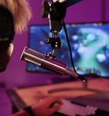 A person wearing headphones is seated in front of a computer screen, engaged in a video game. A microphone is positioned prominently in front of them, suggesting a streaming or recording setup. The background is illuminated with soft purple lighting, creating a cozy and immersive atmosphere.