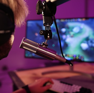 A person wearing headphones is seated in front of a computer screen, engaged in a video game. A microphone is positioned prominently in front of them, suggesting a streaming or recording setup. The background is illuminated with soft purple lighting, creating a cozy and immersive atmosphere.