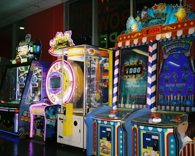 A row of brightly colored arcade game machines is arranged in a line against the wall. Each machine features vibrant lights and designs, with themes ranging from claw games to game show-inspired setups. The area has a playful and inviting atmosphere, suitable for entertainment or amusement centers.