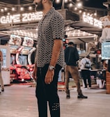 A person stands in a lively arcade setting at night, surrounded by string lights and various arcade game machines. The scene is bustling with activity as other people are visible in the background, engaging with the games or walking by. The signage reads 'Game Zone', indicating the entertainment venue.