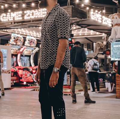 A person stands in a lively arcade setting at night, surrounded by string lights and various arcade game machines. The scene is bustling with activity as other people are visible in the background, engaging with the games or walking by. The signage reads 'Game Zone', indicating the entertainment venue.