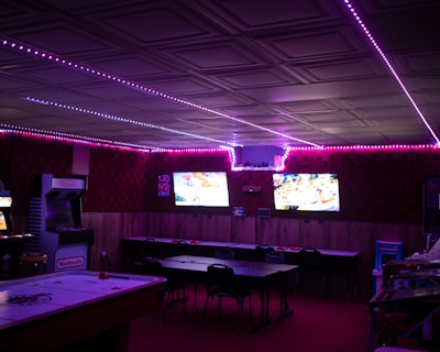 A dimly lit room with vibrant purple LED strip lights on the ceiling, featuring multiple gaming stations. There are two large screens showing video games, along with various arcade machines, including a Nintendo arcade cabinet. An air hockey table is positioned in the foreground.