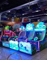 A brightly lit arcade filled with colorful gaming machines sits under ceiling lights. Neon lights illuminate various game stations, creating a playful and vibrant environment. A few red chairs are scattered around the reflective, tiled floor, and a person is observing one of the machines. The atmosphere is lively and cheerful, featuring games with cartoonish designs and engaging themes.