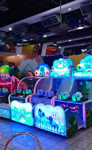 A brightly lit arcade filled with colorful gaming machines sits under ceiling lights. Neon lights illuminate various game stations, creating a playful and vibrant environment. A few red chairs are scattered around the reflective, tiled floor, and a person is observing one of the machines. The atmosphere is lively and cheerful, featuring games with cartoonish designs and engaging themes.