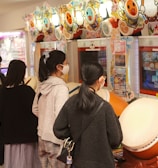Several people are engaged with arcade games, focusing on rhythm games with large drum controllers. The players appear focused and are wearing masks, indicating a casual, indoor setting in an arcade filled with vibrant colors.