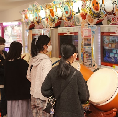 Several people are engaged with arcade games, focusing on rhythm games with large drum controllers. The players appear focused and are wearing masks, indicating a casual, indoor setting in an arcade filled with vibrant colors.