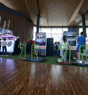 A vibrant arcade setting with several gaming machines lined up, including dance and rhythm game setups. People are engaged with the games, absorbed in their activities. The flooring is wooden, and the ceiling has a geometric design. Natural light filters through large windows, creating a lively atmosphere.